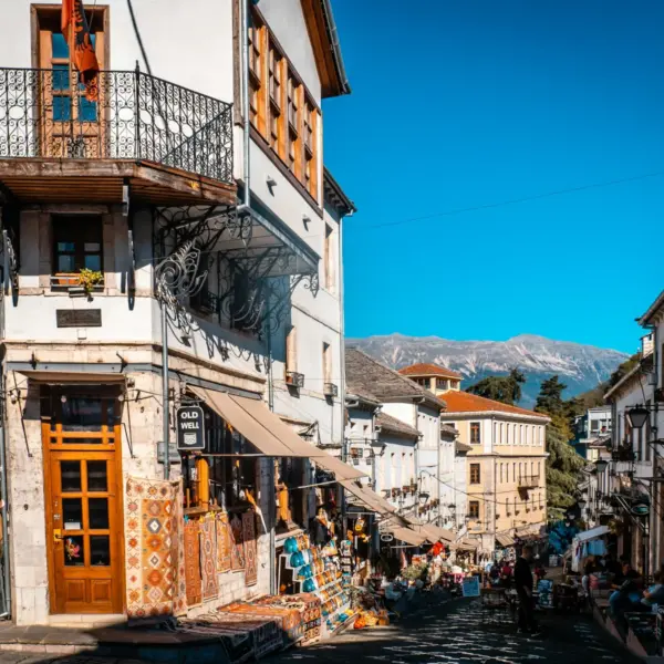 Historic street in Albania lined with traditional buildings, shops, and mountains in the distance