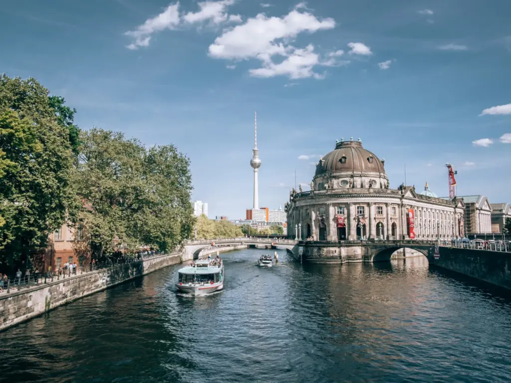 A sightseeing boat travels along the Spree River past Berlin's historic Bode Museum, with the Berlin TV Tower rising in the background.